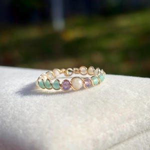 Peut inclure: Une bague dorée délicate ornée de petites pierres précieuses colorées. La conception de la bague incorpore des pierres vert clair, violettes et blanches, tissées ensemble avec du fil d'or. La bague est posée sur un fond blanc doux.