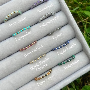 Könnte beinhalten: Nahaufnahme einer Schmuckschatulle mit zehn Edelsteinringen. Die Ringe sind aus Gold und haben verschiedene Edelsteine, darunter Amethyst, Amazonit, Türkis, Labradorit, roter Granat, Pyrit, schwarzer Rutilquarz, Lapislazuli, Tigerauge und Malachit.