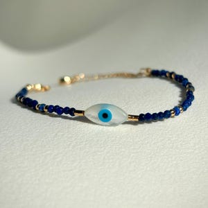 Puede incluir: Un brazalete de cuentas azules y doradas con un dije de ojo turco blanco. El dije tiene un ojo azul en el centro.
