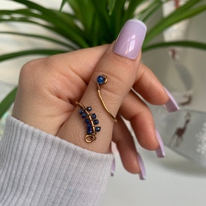 Op de afbeelding: Een gouden ring met een ontwerp van blauwe edelstenen. De ring is om de duim gewikkeld en heeft bovenaan een kleine blauwe edelsteen.