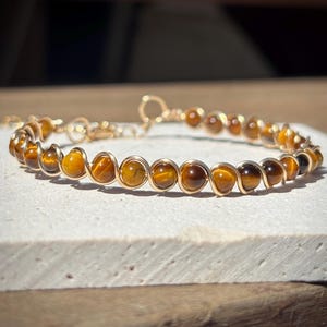 Könnte beinhalten: Ein goldfarbenes, drahtgewickeltes Armband mit Tigeraugenperlen. Das Armband liegt auf einer weißen Oberfläche.