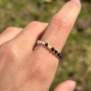 May include: A gold-colored ring with a series of small, round, white and black stones. The ring is on a finger, with a blurred green background.