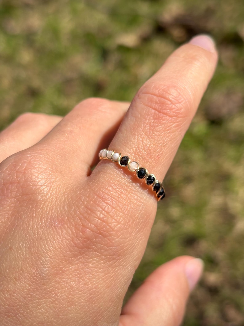 May include: A gold-colored ring with a series of small, round, white and black stones. The ring is on a finger, with a blurred green background.
