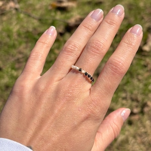 May include: A gold-colored ring with a series of small, black gemstones and white beads. The ring is worn on a hand with a light-colored manicure. The background is blurred, with green and brown tones.