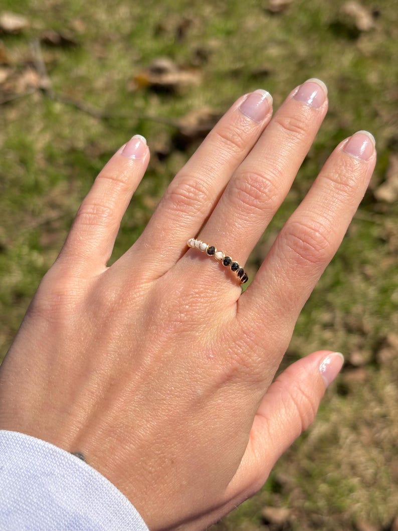 May include: A gold-colored ring with a series of small, black gemstones and white beads. The ring is worn on a hand with a light-colored manicure. The background is blurred, with green and brown tones.
