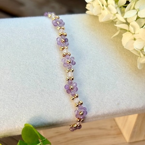 Könnte beinhalten: Ein zartes Armband aus kleinen lila und weißen Perlen, durchsetzt mit goldenen Perlen. Das Armband liegt auf einer weißen Oberfläche.