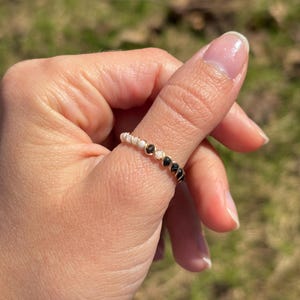 May include: A gold-toned ring adorned with alternating white and black beads. The ring is worn on a finger, showcasing its delicate design and contrasting colors. The beads are evenly spaced around the band.