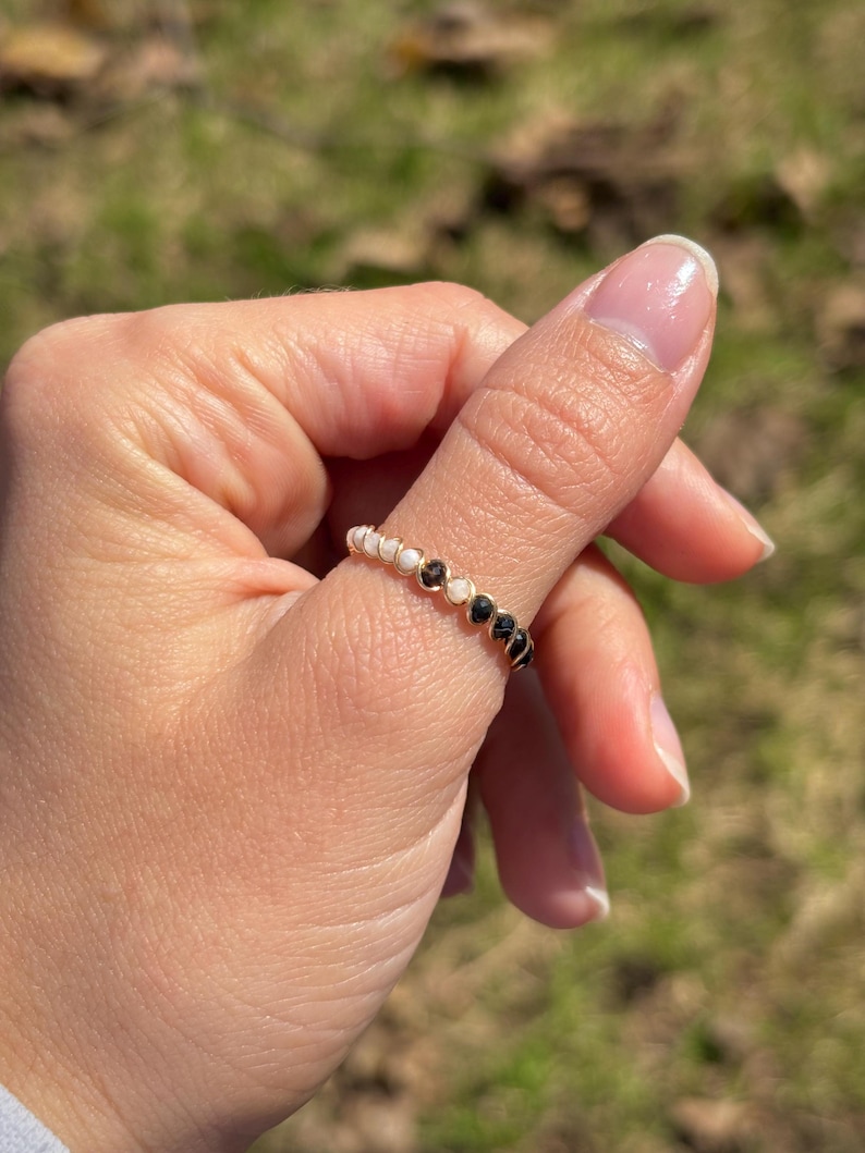 May include: A gold-toned ring adorned with alternating white and black beads. The ring is worn on a finger, showcasing its delicate design and contrasting colors. The beads are evenly spaced around the band.