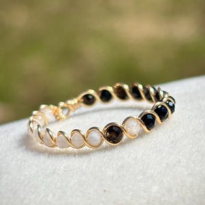 May include: A gold-coloured wire-wrapped ring with alternating small, round gemstones. The gemstones are white, black, and brown. The ring is set against a soft, blurred background.