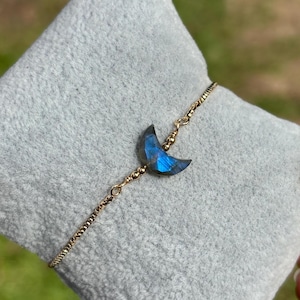 May include: A gold-coloured chain bracelet with a crescent moon pendant. The moon pendant is a faceted, blue gemstone. The bracelet is resting on a grey surface, with a blurred green background.