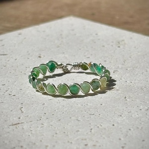 Könnte beinhalten: Ein Ring aus Silberdraht mit grünen Edelsteinen. Der Ring ist aus kleinen, runden, grünen Steinen gefertigt, die in einen Silberdraht gefasst sind.