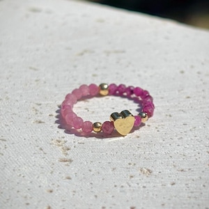 May include: A beaded ring with a colour gradient of pink gemstones, from light to deep magenta. The ring features gold-coloured beads and a gold heart charm, creating a delicate and romantic aesthetic.
