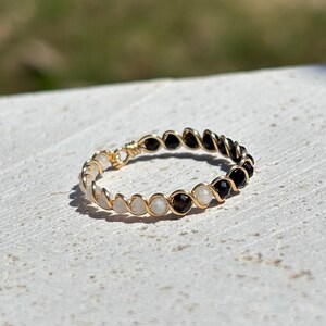 May include: A gold-toned ring with alternating black, white, and brown beads. The beads are strung together with gold wire, creating a delicate and elegant design. The ring is set against a neutral background.