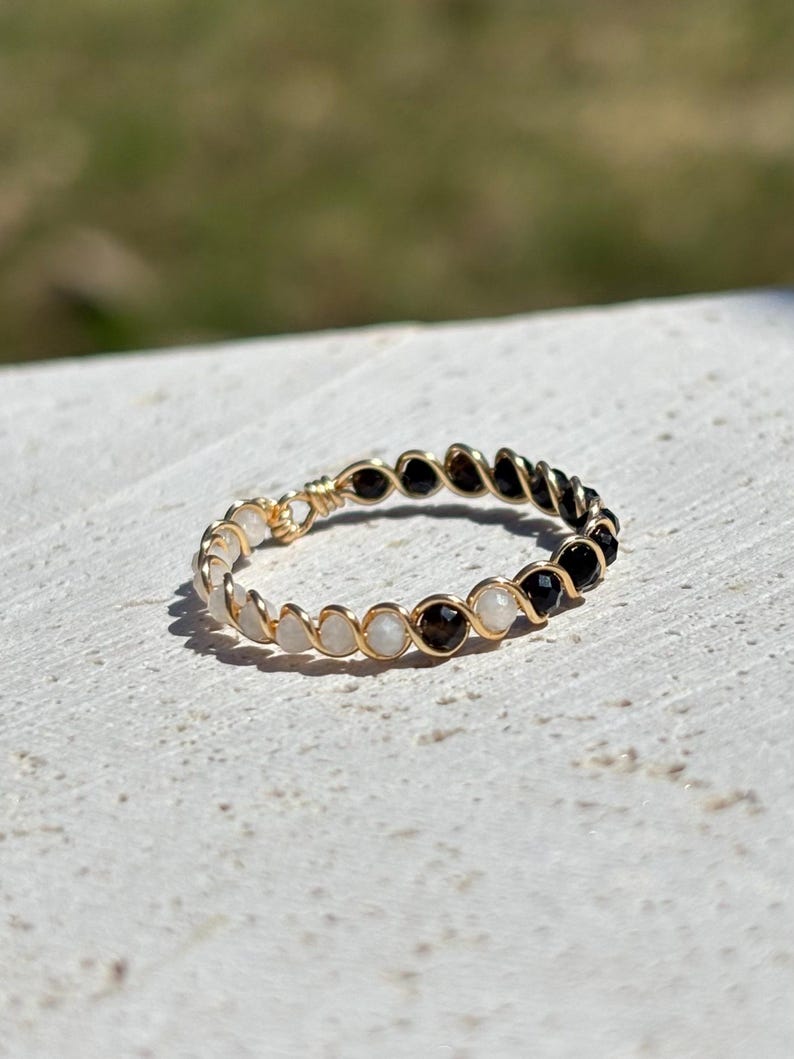 May include: A gold-toned ring with alternating black, white, and brown beads. The beads are strung together with gold wire, creating a delicate and elegant design. The ring is set against a neutral background.