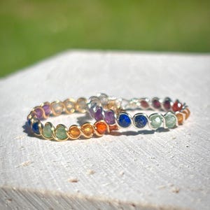 May include: Two rings with colorful gemstones. One ring has a gold band and the other has a silver band. The gemstones are arranged in a rainbow pattern, with colors including blue, green, yellow, orange, and purple.