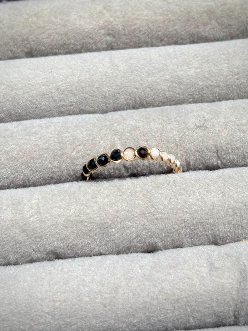 May include: A gold-colored ring with alternating black and white gemstones. The ring is set against a gray, textured background, highlighting the contrast between the ring's metallic sheen and the gemstones.