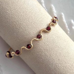 May include: A gold-coloured wire bracelet with a wavy design, featuring several round, deep red gemstones. The bracelet is wrapped around a beige fabric, showcasing the jewellery's intricate details and colour contrast.