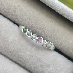 May include: A silver wire wrapped ring with a row of colourful gemstones. The stones are green, blue, purple, and clear.