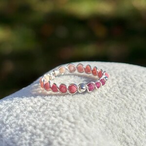 Peut inclure: Une bague faite à la main avec un fil d'argent, ornée de petites pierres précieuses rondes roses et pêche. Les pierres sont étroitement enroulées autour de la bande, créant un motif continu. La bague est posée sur un fond blanc doux.