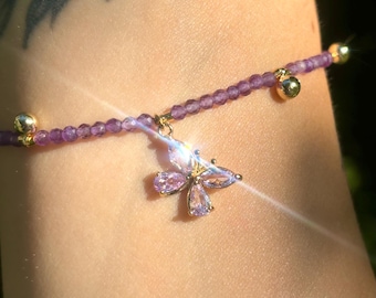 May include: A purple beaded bracelet with a gold butterfly charm. The butterfly has clear and pink rhinestones.