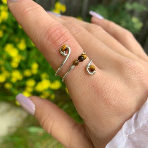 May include: A silver wrap ring with a spiral design. The ring has small, round, brown tiger eye beads. The ring is worn on a finger, with a blurred background of yellow flowers and greenery.