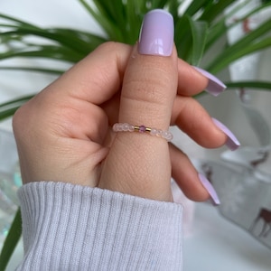Peut inclure: Une délicate bague composée de petites perles blanches avec un accent de pierre précieuse rose. La bague est portée sur un doigt avec un vernis à ongles violet clair.