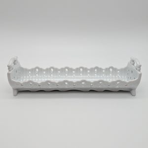 Puede incluir: Una fuente rectangular de cerámica blanca con un borde calado y festoneado. La fuente tiene dos asas pequeñas y descansa sobre cuatro patas pequeñas. El diseño es sencillo y elegante, adecuado para servir aperitivos o artículos pequeños.