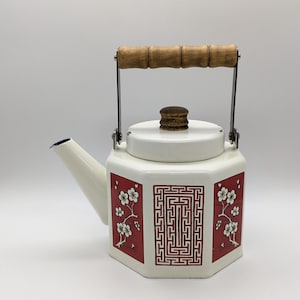 Könnte beinhalten: Weiße Emaille-Teekanne mit Holzgriff und Deckelknopf. Die Teekanne hat rote Paneele mit weißen Blumen- und geometrischen Designs. Der Ausguss ist blau. Ein Vintage-Küchenartikel.