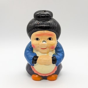 Puede incluir: Figura de cerámica de una mujer sentada con moño negro, chaqueta azul y delantal. La figura tiene una cara sonriente con mejillas rosadas y zapatos naranjas. La figura es un objeto decorativo.