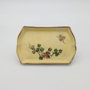 May include: A rectangular ceramic tray with a pale yellow base and a gold-painted rim. The center features a hand-painted botanical illustration of a branch with green leaves and small reddish-brown fruits, plus a small leaf sprig above.
