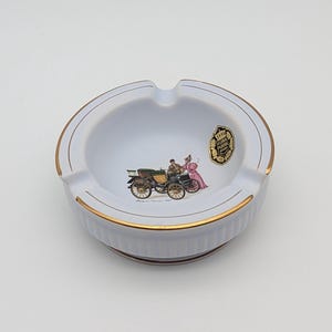 May include: White ceramic ashtray with gold trim and a vintage car illustration. The ashtray features a circular design with a recessed area for holding cigarettes. A gold emblem is visible on the right side of the ashtray.