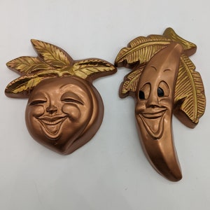 Könnte beinhalten: Zwei vintage Wandplaketten in Form eines Pfirsichs und einer Banane. Beide sind in Kupferfarbe mit goldenen Akzenten bemalt und haben lächelnde Gesichter.