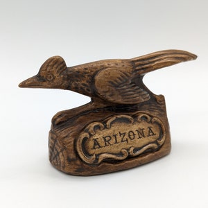 May include: A brown ceramic roadrunner figurine with a carved wooden base. The base has a carved sign that reads "ARIZONA".