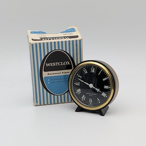 May include: A vintage Westclox keywound alarm clock with a black face, gold-tone bezel, and black base. The clock is next to its original striped blue and white box, which features the Westclox logo and product information.