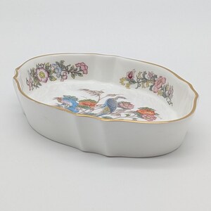 May include: White oval serving dish with a scalloped edge and gold trim. The dish features a colourful floral and bird design in the centre and along the rim. A decorative piece for serving or display.