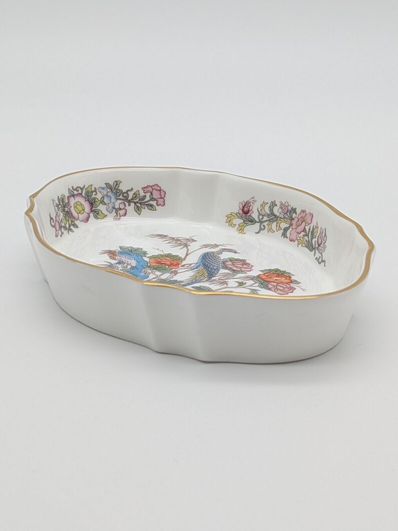 May include: White oval serving dish with a scalloped edge and gold trim. The dish features a colourful floral and bird design in the centre and along the rim. A decorative piece for serving or display.