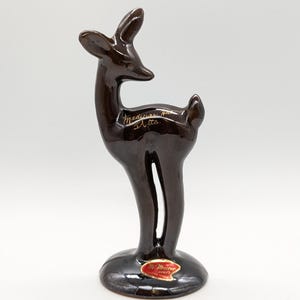May include: A brown ceramic deer figurine with a glossy finish. The deer is standing on a round base and has a slender body with a long neck and small ears. The text "Medicine Hat Alta" is written on the side. A red sticker with gold text is on the base.