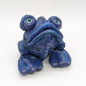 Könnte beinhalten: Eine blaue Keramik-Froschfigur mit großen, runden Augen und einer strukturierten Oberfläche. Der Frosch hat einen breiten Mund und scheint mit eingezogenen Beinen zu sitzen.