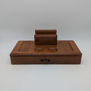 May include: A wooden desk organiser with a drawer and a built-in phone or tablet stand. The organiser is a warm brown colour and has two circular cutouts. A small, decorative handle is on the drawer.