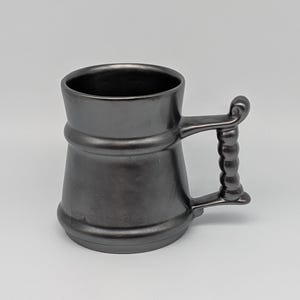May include: A dark grey ceramic beer mug with a handle. The mug has a wide base, a narrower middle section, and a flared top. The handle features a series of vertical ridges and a rounded top.