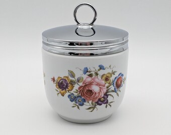 Pode incluir: Um pote de cerâmica branca com uma tampa de metal prateado brilhante com uma alça circular. O corpo do pote é decorado com um padrão floral colorido que consiste em uma rosa rosa central cercada por flores menores azuis, amarelas e roxas.