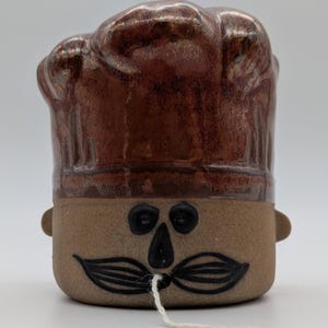 Könnte beinhalten: Eine Keramikfigur in Form eines Kochs mit einer hohen, metallisch braunen Mütze. Die Basis ist ein beiges, zylindrisches Gesicht mit schwarz bemalten Augen, einer Nase und einem Schnurrbart. Ein weißer Faden hängt aus der Mitte des Schnurrbarts.