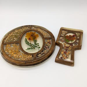 May include: A set of two round, wooden coasters with a clear resin top. The coasters feature a variety of dried flowers and seeds embedded in the resin. The coasters are perfect for protecting surfaces from heat and scratches.