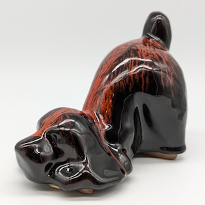 Puede incluir: Una figurita de perro de cerámica con un esmalte negro y rojo. El perro está en una pose juguetona, con la cabeza hacia abajo y la cola hacia arriba.
