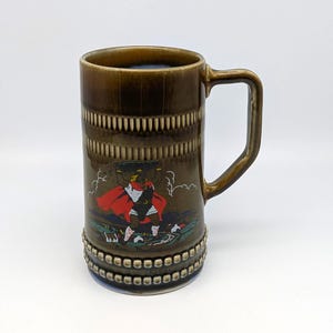 Puede incluir: Una jarra de cerveza de cerámica marrón con asa. La jarra presenta una banda decorativa cerca de la parte superior e inferior, y una imagen central de una figura con capa roja. La jarra mide aproximadamente 15 cm de alto.