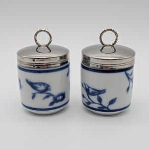 May include: Two cylindrical white porcelain jars featuring blue painted bird and branch motifs. Each jar is topped with a polished silver-toned metal lid that includes a circular ring handle at the centre for easy lifting and access.