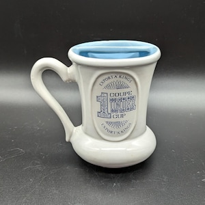 May include: White ceramic mug with a blue interior and handle. The mug features the text "EXPORT'A KINGS" and "COUPE UNDER CUP" in blue lettering. The mug has a unique shape and a rounded base.