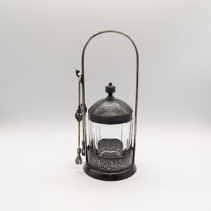 May include: A silver and glass jar with an ornate design and a hinged lid. The jar has a handle and a small spoon attached to the side.