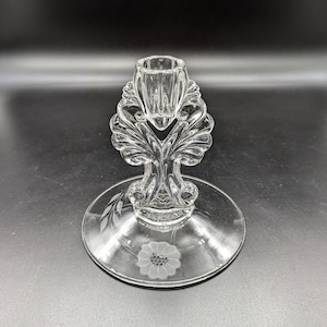 Può includere: Un portacandele in vetro trasparente con un design floreale. Il portacandele ha una base larga e uno stelo alto e sottile con una coppa in cima per tenere una candela.