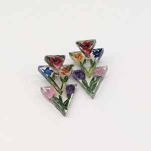 May include: A pair of clear resin earrings with a triangular design. Each earring features colorful miniature flowers in red, yellow, blue, purple, and pink, with green stems and leaves. The earrings are set against a white background.